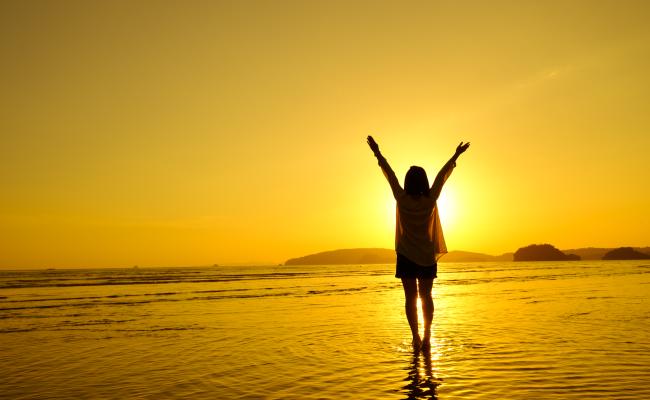 Image of a woman on the beach with arms raised to greet the rising sun