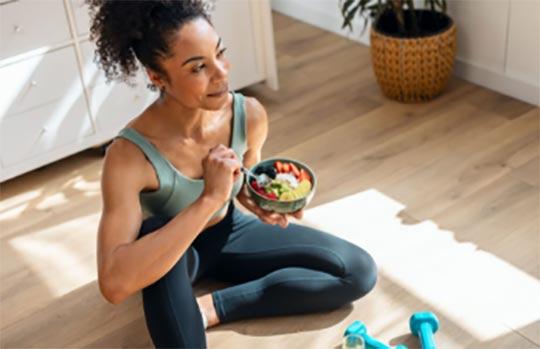 A woman in a workout outfit sitting on the floor with dumbells in front of her and eating healthy.