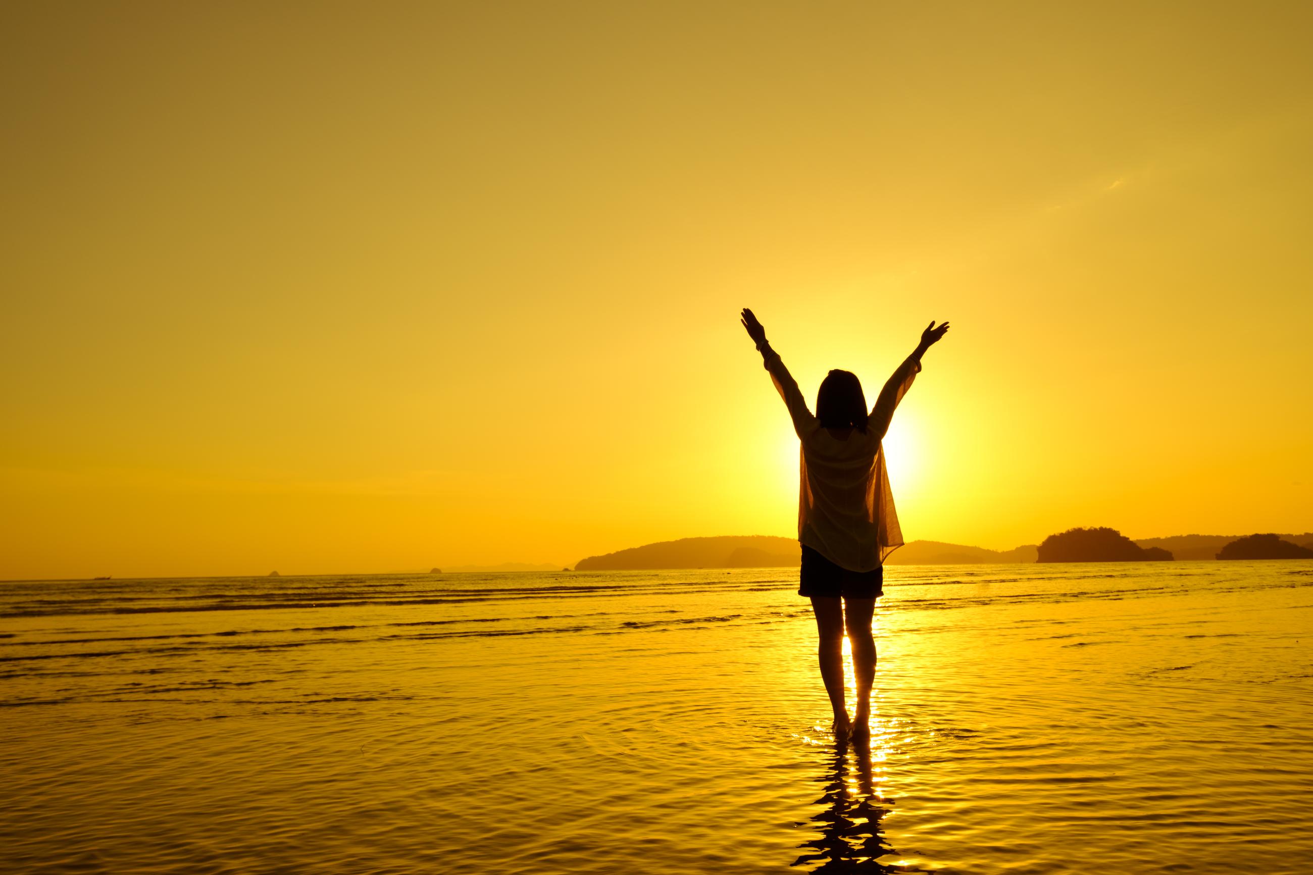 Image of a woman on the beach with arms raised to greet the rising sun