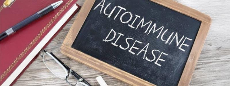 Notebook with a pen laying on it and glasses on the surface beside a small chalkboard with "Autoimmune Disease" written in chalk upon it.